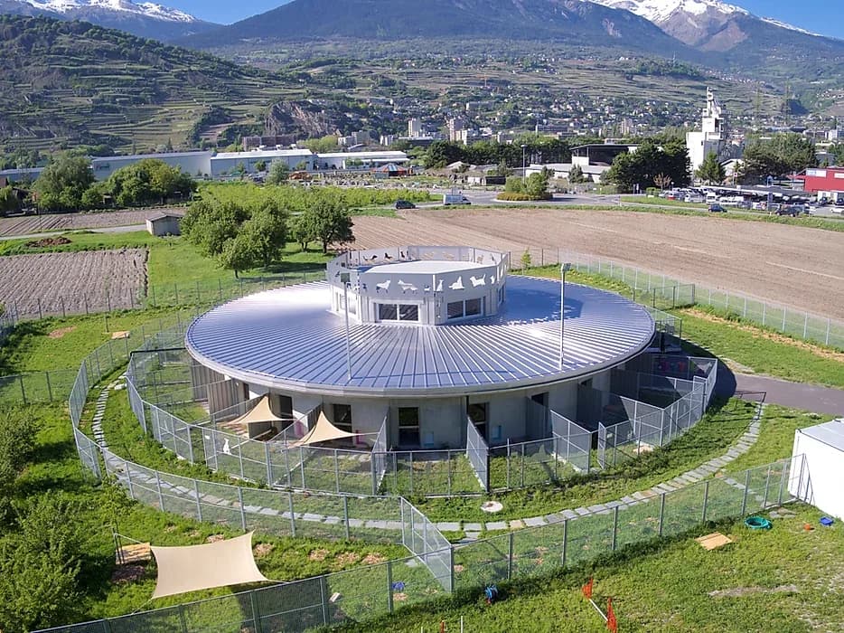 Vue aérienne de la pension : bâtiment rond entouré de parcs clôturés, avec Sion et le coteau en arrière-plan